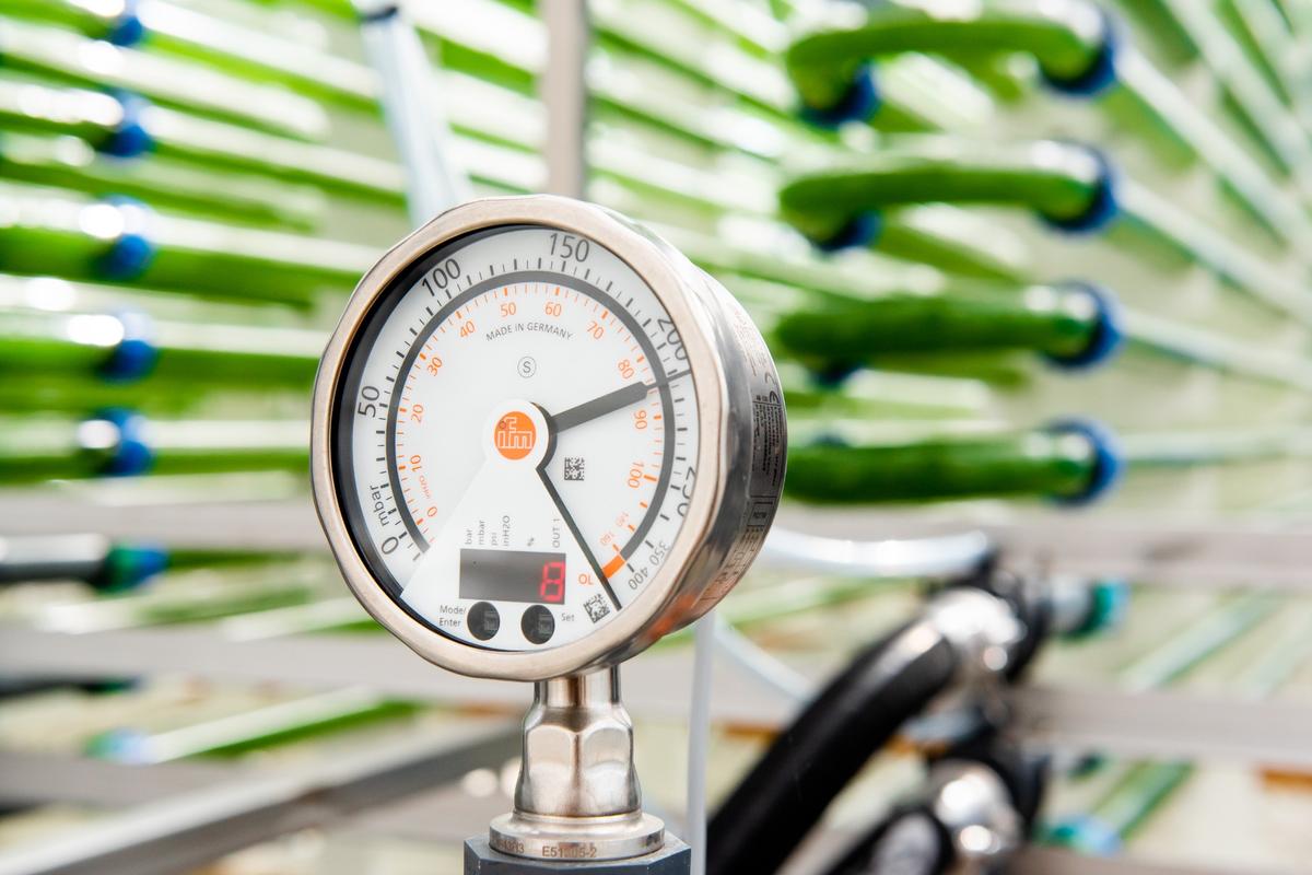 Close-up of a pressure gauge with needle and digital readout for monitoring, against a blurred background of green tubes.